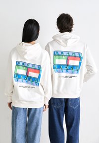 Dvaja ľudia v bielych mikinách s farebnými logami Tommy Jeans na chrbte stoja jeden vedľa druhého na jednoduchom pozadí.