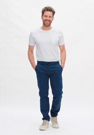 Weißes T-Shirt mit kurzen Ärmeln und Tasche, kombiniert mit dunkelblauen Hosen. Das Modell trägt helle Sportschuhe. Schlichte, lässige Passform.