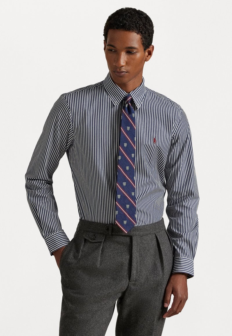 Chemise à rayures bleu marine et blanche avec un col classique, accompagnée d'une cravate bleu marine ornée d'un motif de blason et de pantalons plissés gris.