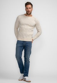 Beiger Kabelstrickpullover mit rundem Halsausschnitt, kombiniert mit blauen Jeans und weißen Sneakers mit schwarzen Akzenten. Schlichtes strukturiertes Design.