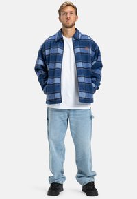 Blue fleece jacket with a checkered pattern, featuring a collar and zip closure, worn over a white t-shirt and paired with light blue jeans.