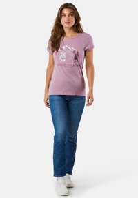 Kortærmet, rundhalset, lyse lilla t-shirt med et grafik af et bjerg og en bjørn med træer. Bæres med mørkeblå jeans og hvide sneakers.