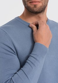 Lichtblauwe sweater met een geribde hals en mouwen, toont een soepele textuur. Een hand houdt de kraag lichtjes vast.