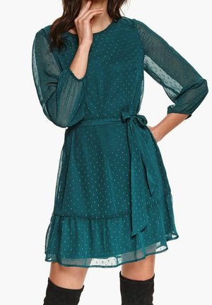 Donna che indossa un vestito verde acqua a maniche lunghe con pois testurizzati, vita annodata e orlo con balze abbinato a stivali neri al ginocchio.