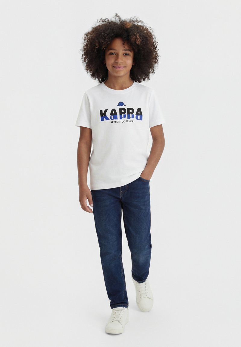 Kind mit lockigem Haar geht, trägt ein weißes Kappa-T-Shirt, dunkelblaue Jeans und weiße Sneakers vor einem einfarbigen hellen Hintergrund.