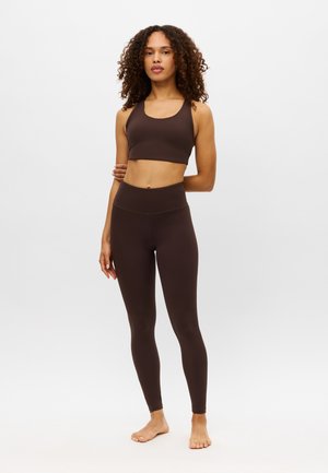 Kvinde med krøllet hår iført en mørkebrun sports-bh og leggings med høj talje, stående barfodet mod en ensfarvet lys baggrund.