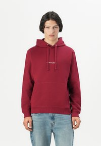 Bordo spalvos megztinis-kapucha su priekiu kišeniu, raišteliu ant galvos ir balta logotipo detalė. Sukombinuotas su šviesiai mėlynomis džinsomis. Pagamintas iš medvilnės mišinio.