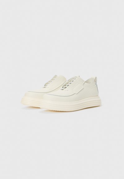 Hvide slip-on sneakers med en tekstureret overflade, der har et syet design og en tyk, rillede sål. Minimalistisk æstetik uden synlige snørebånd.