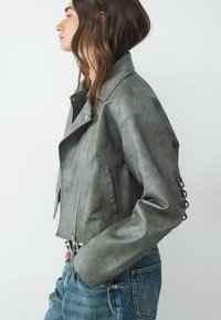 Geknipte grijze leren jas met een ingesneden kraag, ritssluiting en gevlochten details op de mouwen. Gepaard met lichte spijkerbroek.