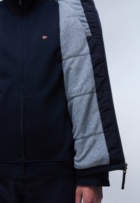 Marineblaue Zip-Jacke mit grauen, gesteppten Ärmeln. Verfügt über ein kleines Logo auf der Brust und gerippte Bündchen. Weicher, strukturierter Stoff.