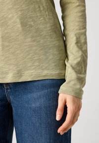 Langermet, lysegrønn topp med teksturert overflate, kombinert med mørkeblå denimjeans. Ermet har et tettsittende håndledddesign.
