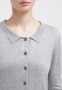 Cardigan grigio a maglia con collettino, dotato di cinque bottoni di colore più scuro. Texture morbida con silhouette aderente.