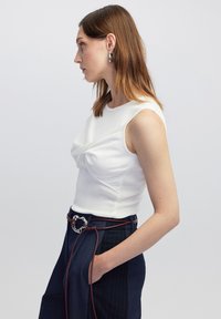Witte mouwloze top met geplooide stof aan de voorkant, gecombineerd met marineblauwe broek met streepjes en een geweven riem met een metalen gesp.