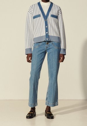 Personne portant un cardigan pied-de-poule blanc et bleu avec des bordures en denim bleu, un jean bleu clair et des mocassins noirs avec des chaînes dorées.