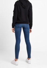 Felpa nera con cappuccio e tasca a marsupio, abbinata a jeans skinny blu e sneakers bianche. I jeans presentano un orlo leggermente sfilacciato.