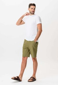 Mand iført hvid t-shirt, olivenfarvede shorts og brune sandaler, stående med den ene hånd i lommen og ser til siden.