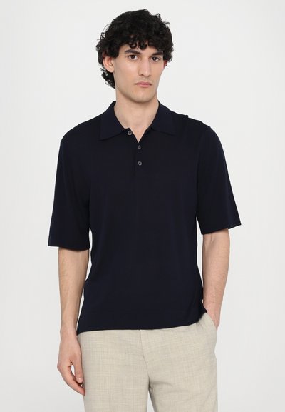 Polo shirt bleu marine à manches courtes avec un col plat, trois boutons et une texture lisse. Porté avec un pantalon beige texturé.