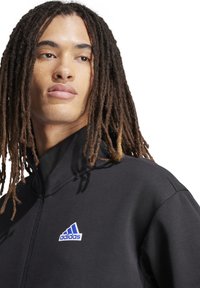 Zwarte Adidas zip-up jacket met hoge kraag, met een blauw logo op de linkerkant van de borst. Gladde stof en aansluitend ontwerp.