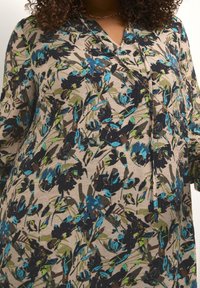 Tunika floreale beige con motivi blu, verdi e neri. Presenta una scollatura a V e maniche lunghe in un tessuto testurizzato.