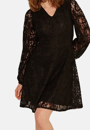 Woman wearing a black lace dress with long sheer sleeves and a V-neckline, standing with one hand on her hip.