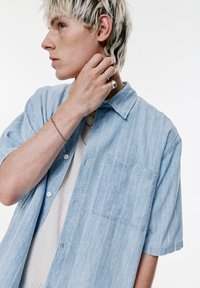 Camicia azzurro chiaro a maniche corte con bottoni, realizzata in materiale testurizzato, con strisce verticali e una tasca sul petto. Indossata sopra un top in maglia beige.