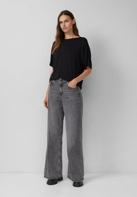 Kobieta stoi z rękami w kieszeniach, ubrana w czarną luźną bluzkę z krótkim rękawem, szare szerokie dżinsy oraz czarne masywne buty na tle szarej ściany.