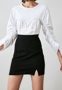 Žena v bílém dlouhém rukávu s nařaseným topem a černou sukní s vysokým pasem a bočním rozparkem, stojící proti jednobarevnému pozadí.