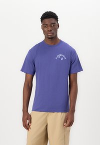 Lilla bomulls T-skjorte med korte ermer og rund hals. Har hvit tekst og grafisk design på øvre venstre brystområde.