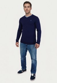 Marineblauwe ribgebreide trui met een V-hals en lichtblauwe accenten, gecombineerd met lichtblauwe spijkerbroeken en donkere sneakers.