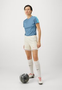 Zila īsām piedurknēm sporta krekls, baltas bikses ar krāsainiem akcentiem, līdz ceļiem baltas zeķes un futbola bumba. Nike zīmols uz krekla, biksēm un zeķēm.