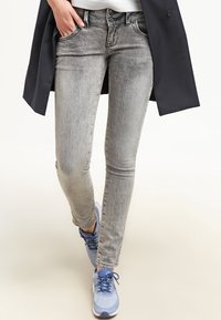 Jean skinny gris avec une finition délavée, design à cinq poches et fermeture à bouton standard, assortis à des chaussures de sport bleues.