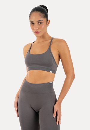 Donna con i capelli raccolti in uno chignon, indossa un reggiseno sportivo grigio e leggings vita alta, in piedi con una mano appoggiata al fianco.