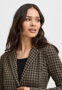 Brauner Hahnentritt-Blazer aus strukturiertem Stoff mit Reverskragen und einem schwarzen Innenfutter. Das Design ist figurbetont und strukturiert.