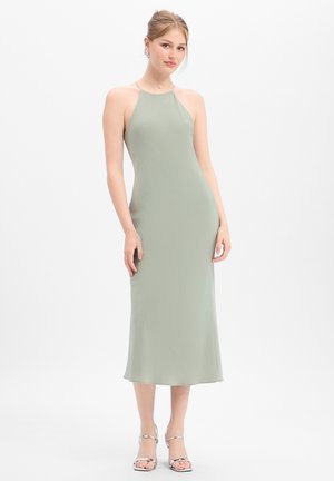 Jeune femme portant une robe midi vert clair sans manches et des talons argentés, debout devant un fond blanc uni.