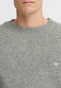 Siv pulover z ribanim pletenjem, ki ima okrogel izrez in majhen bel logotip na levem prsnem delu. Mehka tekstura z oprijetim krojem.