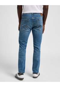 Jeans de denim azul con corte slim, con cinco bolsillos, costura en el bolsillo trasero y un parche de cuero de la marca cerca de la cinturilla. Se combinan con zapatillas blancas.