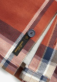 Geruite stof shirt in roest, marineblauw en crèmewit. Heeft een blauwe knop en een label met de tekst "DURABLE GOODS" in zwarte letters.