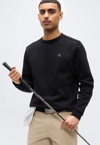 Νεαρός άνδρας με μαύρο φούτερ κρατώντας ένα γκλογκ, με λευκό γάντι γκολφ κρυμμένο στα μπεζ παντελόνια, πάνω σε απλό φόντο.