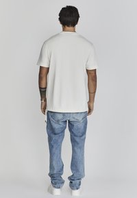 Camisa blanca de manga corta, vaqueros de denim holgados y zapatillas blancas. La camisa tiene un dobladillo discreto y los vaqueros presentan un lavado azul desgastado.