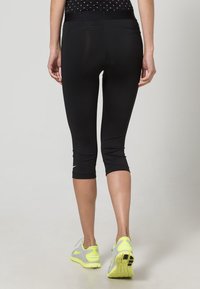 Czarne legginsy capri z szerokim paskiem, dopasowanym krojem i odblaskowym logo Nike. Noszone z szaro-neonowożółtymi butami sportowymi.