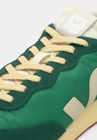 Πράσινο αθλητικό παπούτσι με υφή από σουέντ, μπεζ κορδόνι και λευκή ετικέτα με λογότυπο. Διαθέτει κυματιστό σχέδιο και αντίθετη κρεμ σόλα.