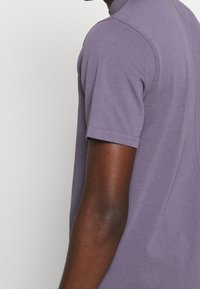 Bras et épaule d'un homme portant un t-shirt violet à manches courtes sur fond clair.