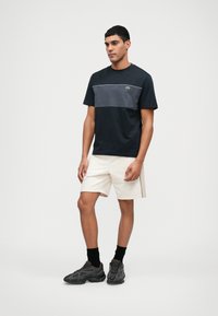 Marine T-shirt met een grijze horizontale streep, ronde hals; gecombineerd met crèmekleurige shorts met zijnaad, gedragen met zwarte sokken en grijze sneakers.