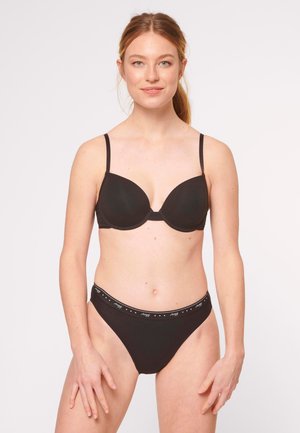 Mujer de piel clara y cabello recogido, vestida con un sujetador negro y ropa interior negra de marca, de pie frente a un fondo claro y liso.