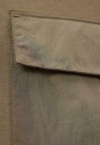 Inquadratura ravvicinata di una tasca in tessuto khaki con patta, che mostra i dettagli delle cuciture e la texture su uno sfondo di un indumento lavorato a maglia.