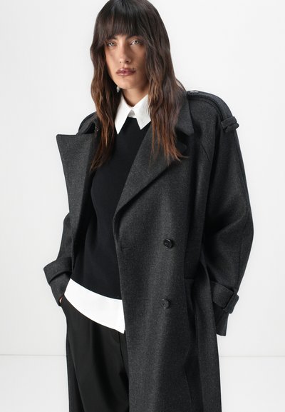 Langer grauer Mantel aus strukturiertem Stoff, mit einem doppelt geknöpften Design, großen Revers und hochgeschlagenen Ärmeln über einem schwarzen Sweater und einem weißen Hemd.