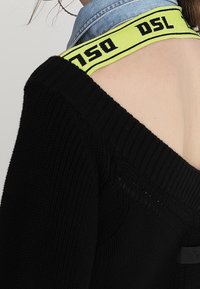 Svart stickad tröja med off-shoulder-design, med en neongul remsa märkt "DSL" och en blå denimkrage.