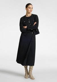 Musta räätälöity takki, jossa on kaksi rintataskua, yhdistettynä virtaavaan mustaan hameeseen ja polviin saakka ulottuviin beigekenkkiin. Sujuva kangas, jolla on rakenteellinen istuvuus.