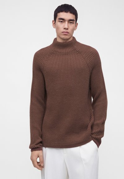 Jeune homme portant un pull à col roulé côtelé marron et un pantalon blanc, debout devant un fond clair uni.