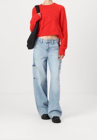 Suéter rojo corto con detalles texturizados y mangas largas, combinado con pantalones cargo de pierna ancha en azul claro con bolsillos laterales. Bolsa de hombro negra.